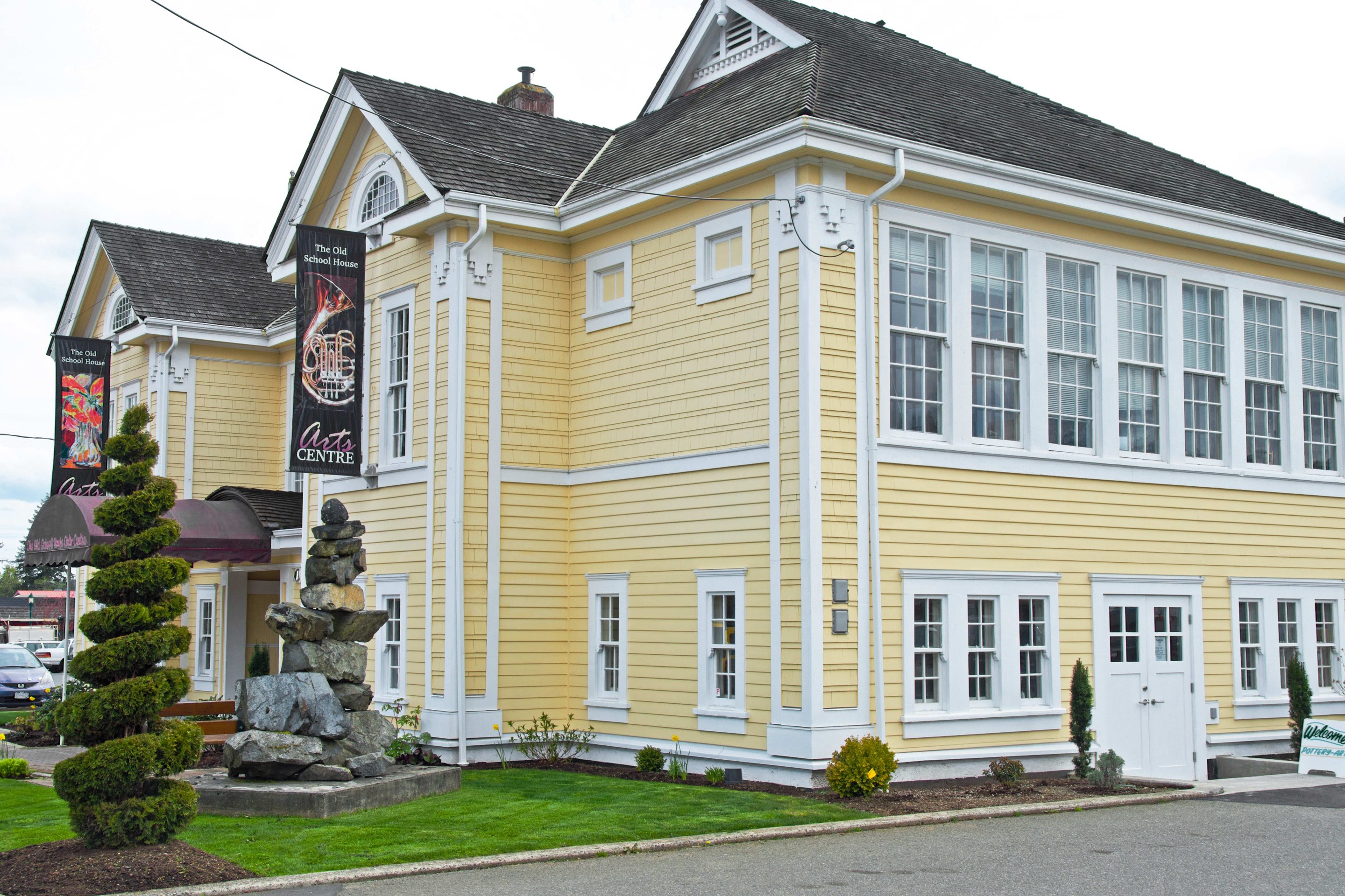 Exterior of The Old School House Arts Centre, a charming yellow heritage building with white trim located in Qualicum Beach, British Columbia. A banner featuring a brass instrument and the words "Arts Centre" hangs on the side. A stacked stone sculpture is displayed on the manicured lawn in front.