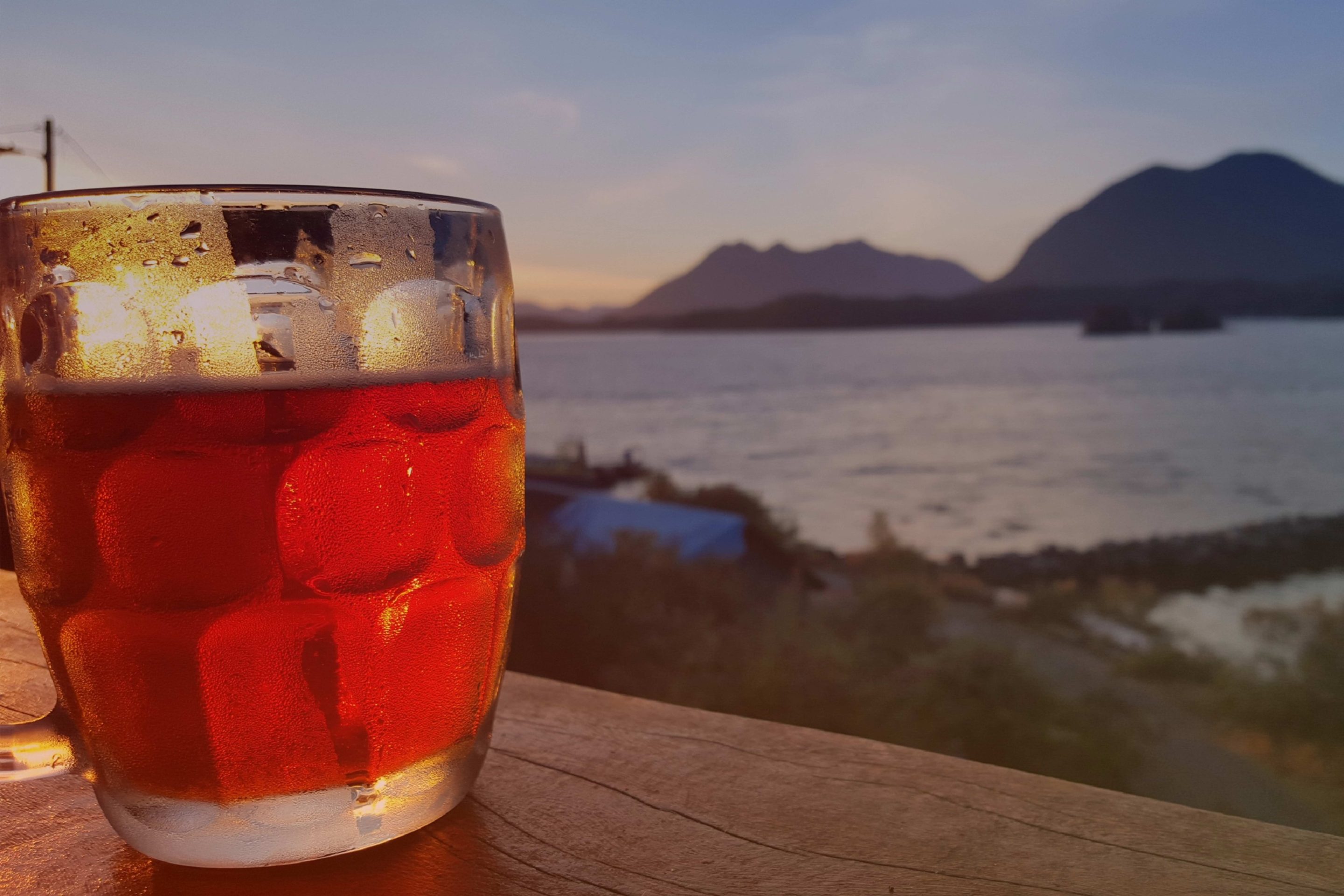 Close-up of a chilled glass of amber-colored beverage with ice, set on a wooden surface. In the background, a scenic view of a calm ocean inlet and mountains at sunset, creating a peaceful and inviting atmosphere.