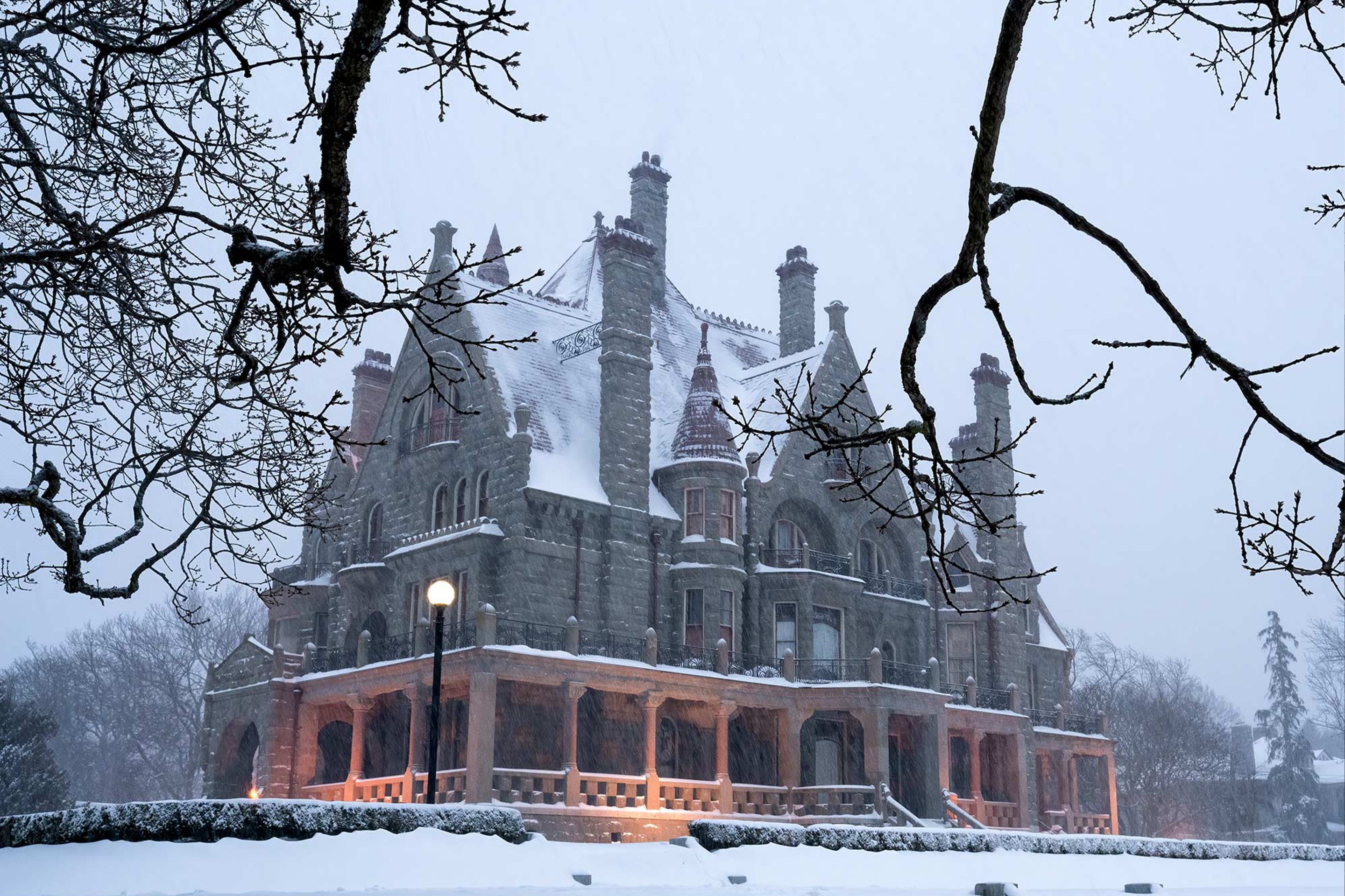 Craigdarroch Castle in Victoria, British Columbia, covered in snow during winter. The historic stone mansion is surrounded by leafless tree branches and soft snow blankets the ground, creating a serene, picturesque scene.