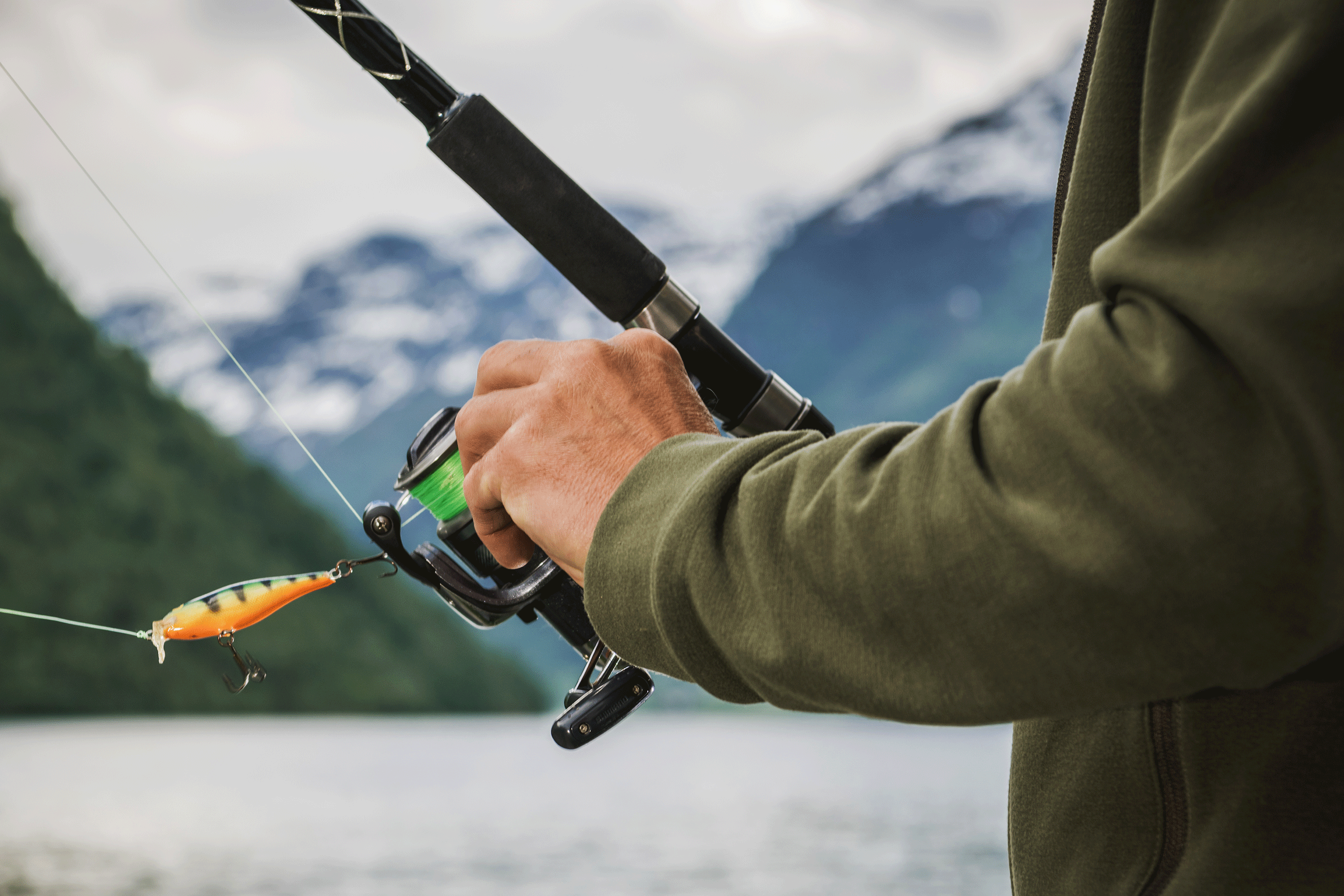 Close-up of a person holding a fishing rod with an orange lure, preparing to cast into a serene lake. Snow-capped mountains and forested hills provide a dramatic backdrop under an overcast sky