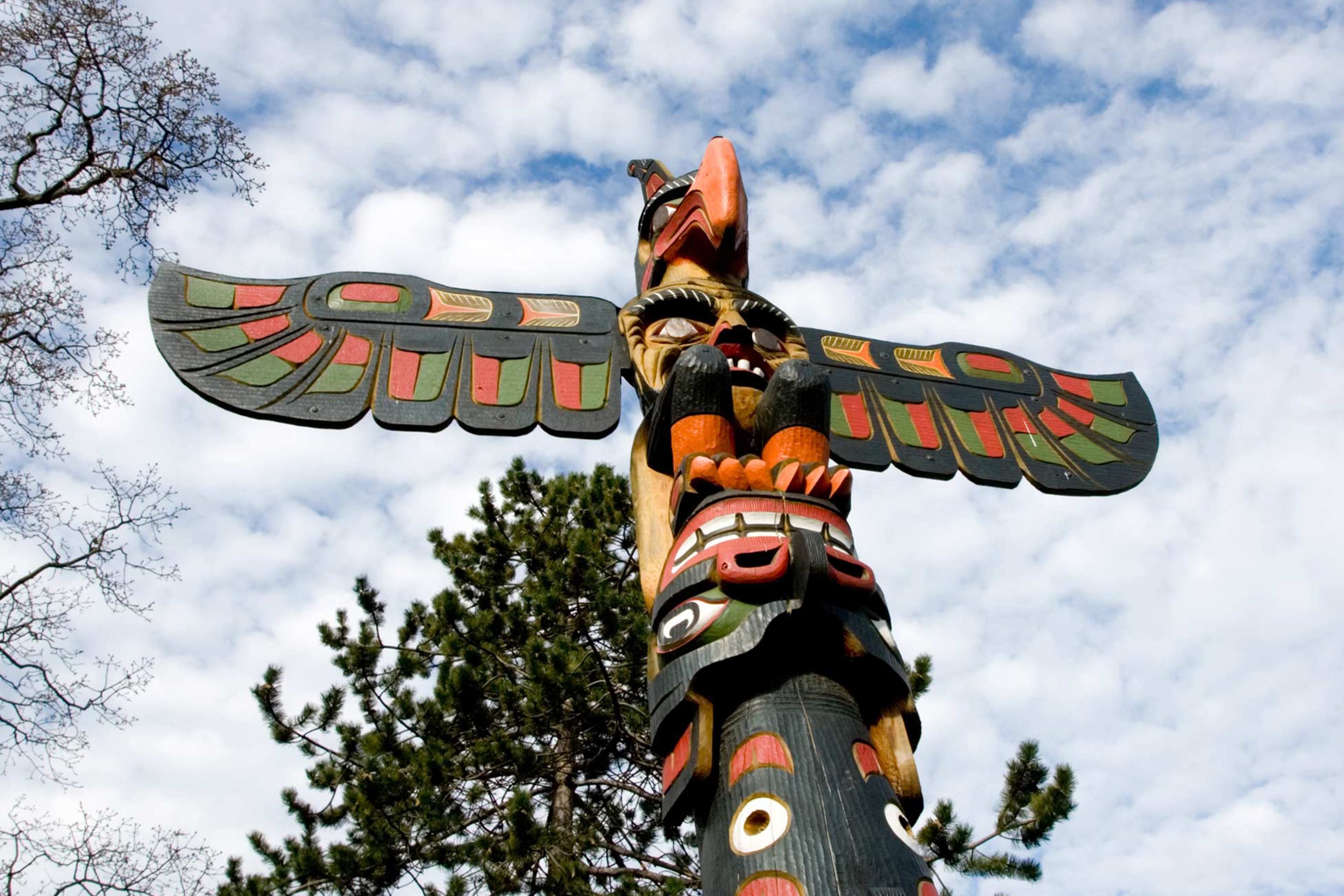Upward view of a brightly painted Indigenous totem pole set against a partly cloudy sky. The carving features a large bird with outstretched wings at the top, surrounded by symbolic animal figures. Trees frame the background, highlighting the totem’s natural setting.