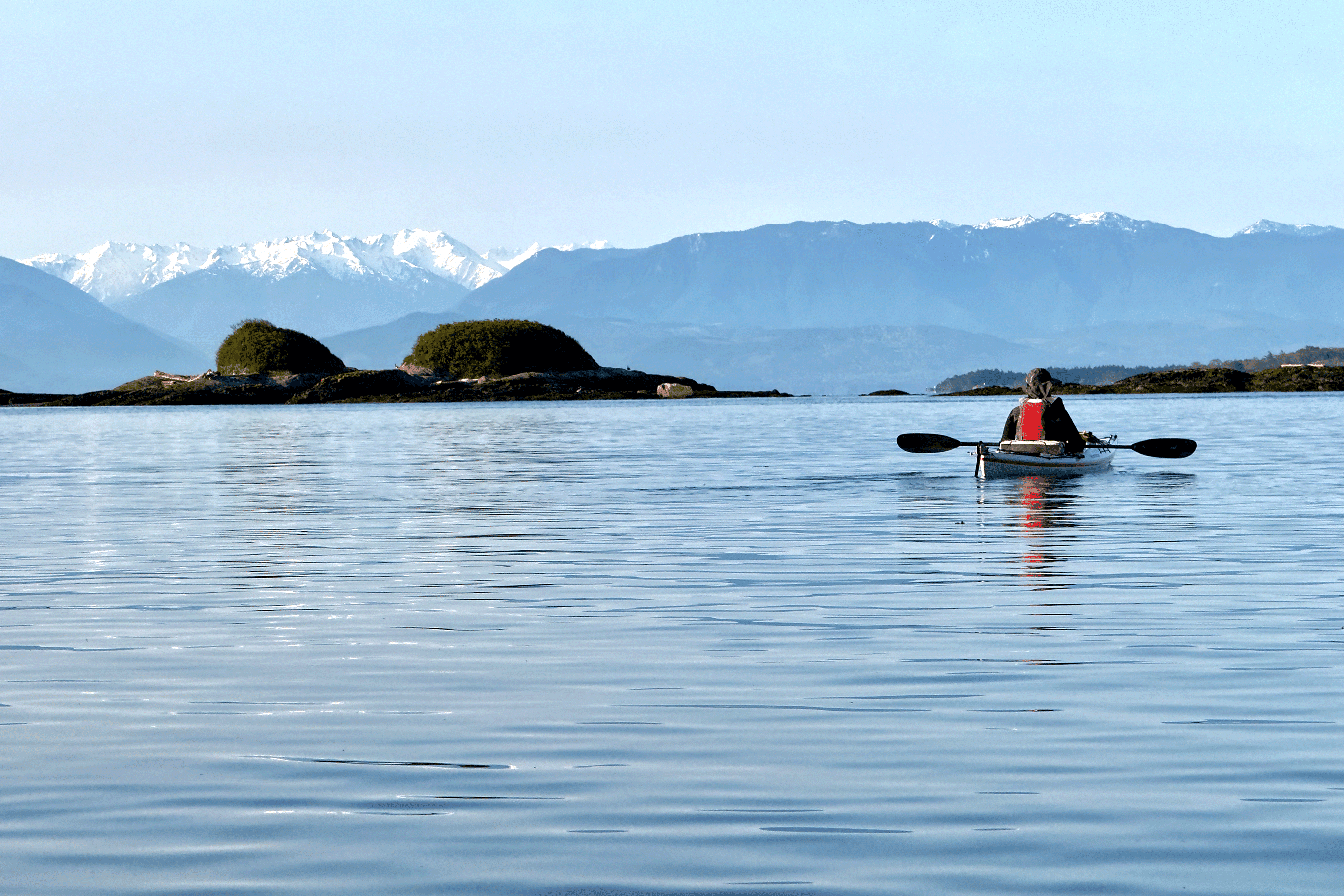A person in a kayak paddles across calm coastal waters, surrounded by small rocky islets. Snow-capped mountains rise in the distance under a clear blue sky, creating a peaceful and scenic outdoor setting.