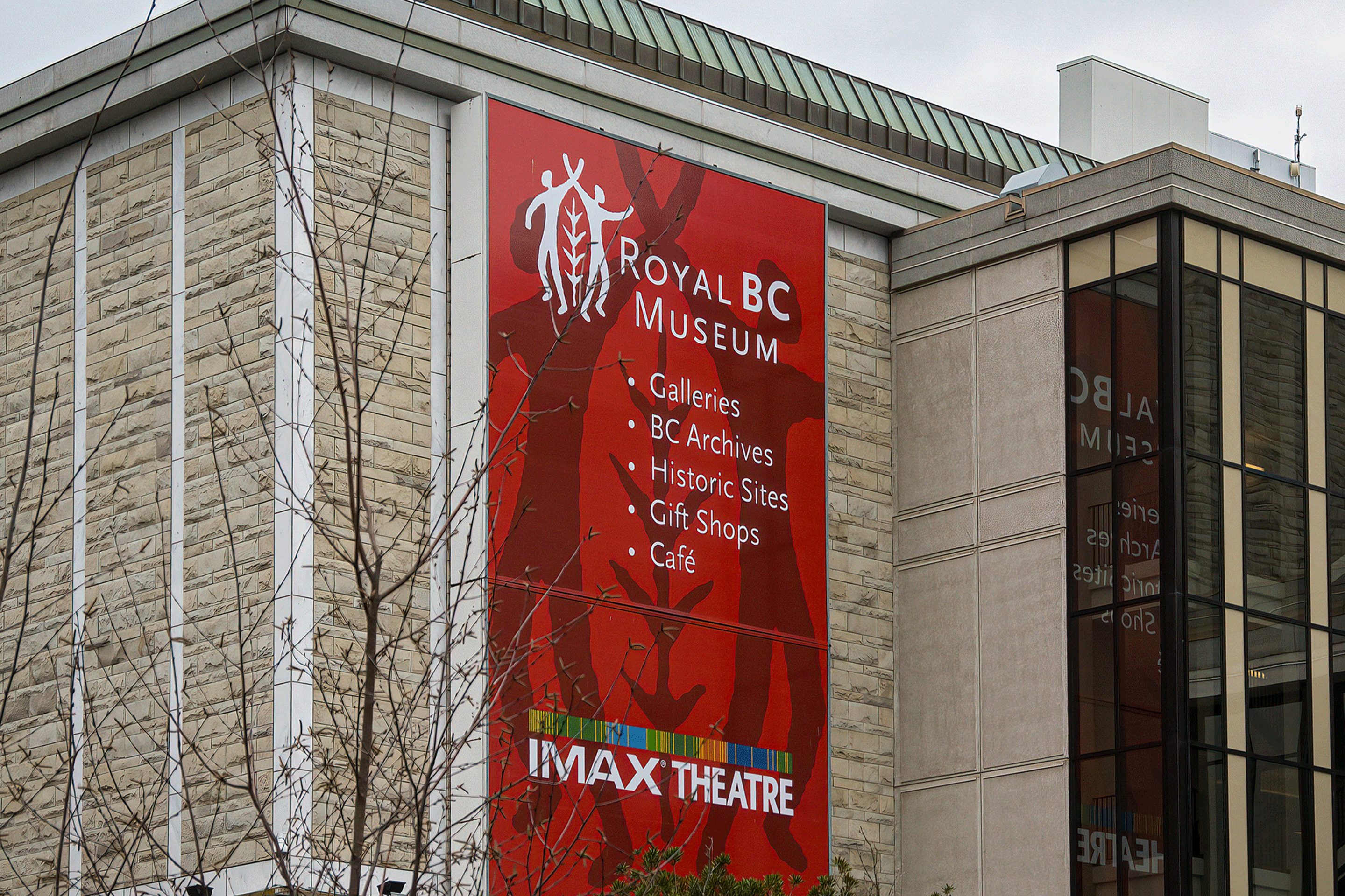 Exterior view of the Royal BC Museum building in Victoria, British Columbia. A large red banner on the stone facade lists museum features including Galleries, BC Archives, Historic Sites, Gift Shops, and Café. The sign also advertises the IMAX Theatre located within the museum.